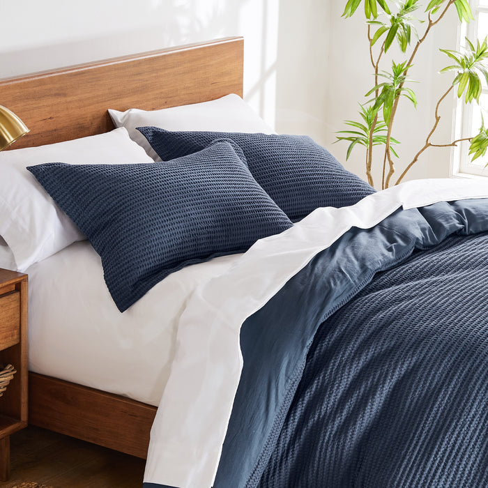 A modern twin bed with a navy waffle-textured comforter set, white sheets, and a wooden headboard, brightened by natural light and greenery.