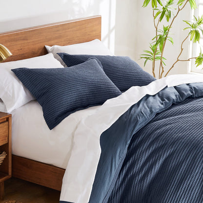 A modern twin bed with a navy waffle-textured comforter set, white sheets, and a wooden headboard, brightened by natural light and greenery.