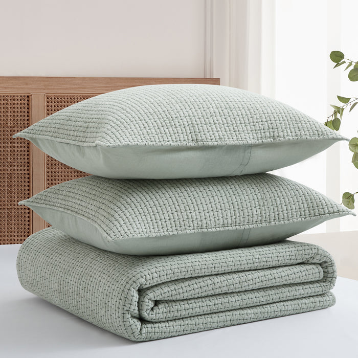 Stack of light green textured pillows and a blanket on a bed with a neutral background